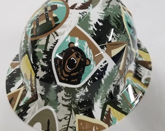 Puede incluir: Un casco blanco con un patrón de bosque verde, marrón y azul. El casco presenta un gráfico de oso y varias ilustraciones con temática de camping.