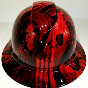 May include: Red hard hat with a skull and playing card graphic. The skull is wearing a hood and holding a playing card with a spade symbol.