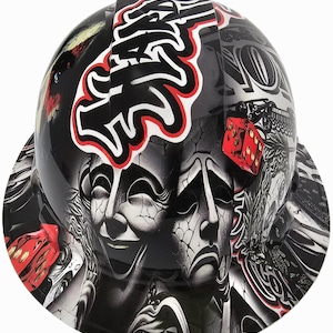 May include: A black hard hat with a detailed graphic design. The design includes a pair of theatrical masks, dice, and stylized text outlined in red and white. The overall theme is a blend of art and industrial safety.