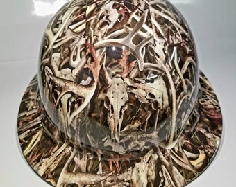 Puede incluir: Un casco de obra con un patrón de camuflaje que presenta cuernos y cráneos. El patrón está en tonos de marrón, blanco y negro.