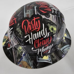 May include: A black hard hat with a money print design. The hat has the words "Dirty Hands, Clean Money" written in red and white on the front.