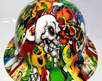May include: A white hard hat with a colorful graphic design featuring skulls, flames, and graffiti. The design is a mix of red, green, yellow, blue, and black.