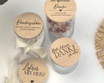 May include: Four glass jars with wooden lids, each with German text. One jar contains dried flowers. The lids have phrases like "Thank you" and "Teacher with Heart". A white ribbon and eucalyptus leaves are also visible.