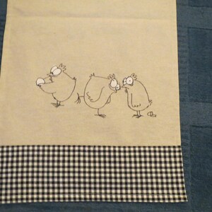 Könnte beinhalten: Ein beiges Küchentuch mit einer humorvollen Illustration von drei Cartoon-Hühnern. Die Hühner haben große Augen und sind mit schwarzer Tinte gezeichnet. Der untere Rand hat ein schwarz-weißes Karomuster.