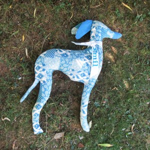 May include: A stuffed fabric dog toy featuring a patchwork pattern of various blue and white floral and paisley prints. The dog has a white ribbon tied around its neck with the name Emil printed in blue letters on the fabric ribbon.