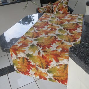 Könnte beinhalten: Ein rechteckiger Tischläufer mit einem Herbstblattmuster in Orange, Gelb und Grün auf weißem Hintergrund. Der Läufer ist über einen Tisch drapiert und zeigt das herbstliche Design. Ein passendes Kissen ist im Hintergrund zu sehen.