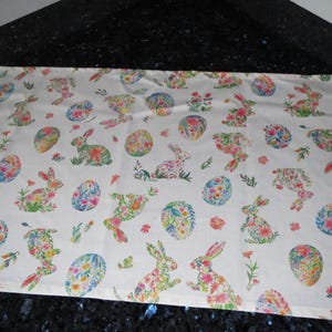 Könnte beinhalten: Weißer, rechteckiger Tischläufer mit einem sich wiederholenden Muster aus floralen Hasen und dekorierten Ostereiern. Die Hasen und Eier sind mit bunten Blumen in Rosa-, Gelb-, Blau- und Grüntönen gefüllt.