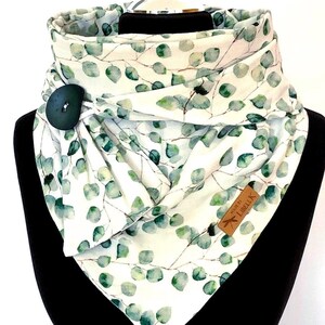 Könnte beinhalten: Weißer Schal mit grünem Eukalyptusblattmuster und großem dunkelgrünen Knopfverschluss. Der Schal ist um einen schwarzen Schaufensterkopf gewickelt. Ein kleines braunes Etikett mit der Aufschrift "Made by Libellk" ist angebracht.