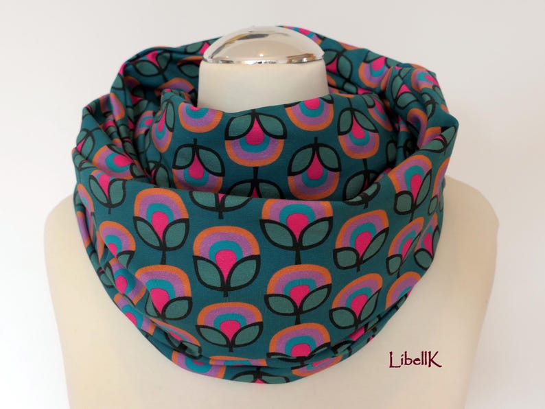 May include: Teal blue infinity scarf with a repeating pattern of pink, orange and purple flowers. The flowers have a retro design with a circle in the centre.