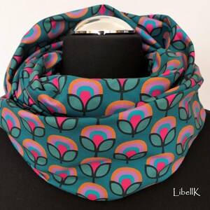 May include: Teal blue infinity scarf with a repeating pattern of pink, orange, and teal flowers. The scarf is folded in half and is lying flat on a black surface.