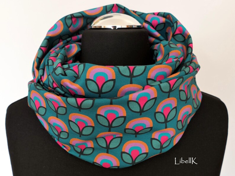 May include: Teal blue infinity scarf with a repeating pattern of pink, orange, and teal flowers. The scarf is folded in half and is lying flat on a black surface.