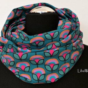 May include: Teal blue infinity scarf with a repeating pattern of pink, orange, and teal flowers. The scarf is folded and displayed on a black mannequin.