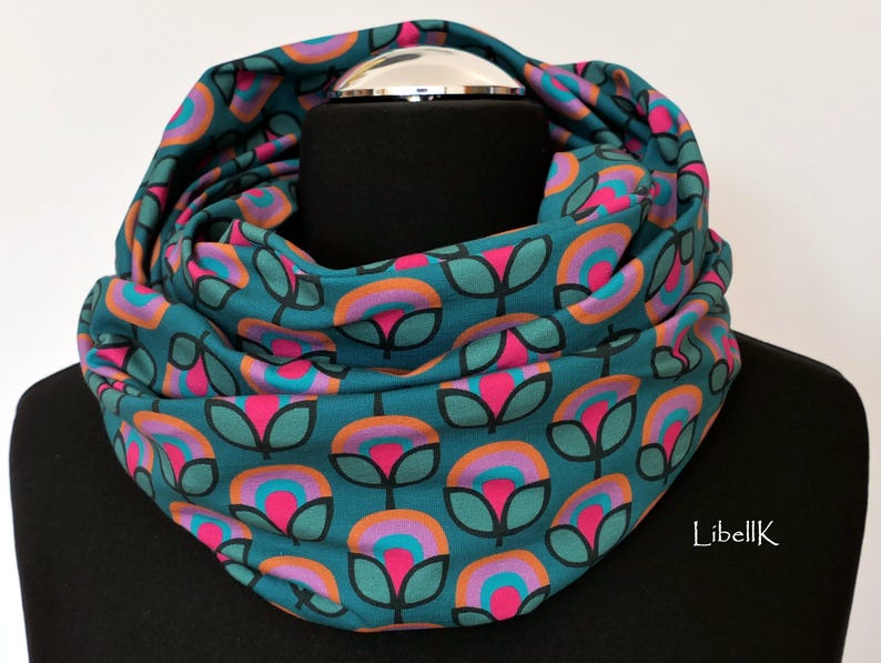 May include: Teal blue infinity scarf with a repeating pattern of pink, orange, and teal flowers. The scarf is folded and displayed on a black mannequin.
