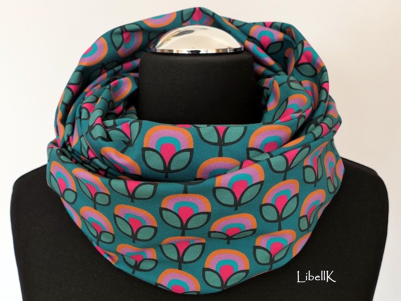 May include: Teal blue infinity scarf with a repeating pattern of pink, orange, and teal flowers. The scarf is folded and displayed on a black mannequin.