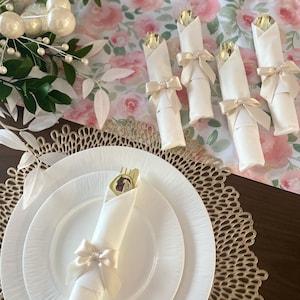 May include: A white dinner plate topped with a white napkin rolled around gold-toned cutlery and secured with a cream-colored satin ribbon bow. Additional rolled napkins with gold cutlery and ribbons are arranged on a floral table runner.