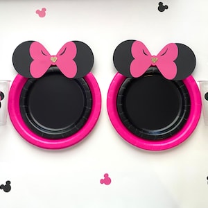 May include: Two black and pink paper plates with a pink bow and black ears on top. The plates are on a white surface with black and pink mouse ears scattered around.