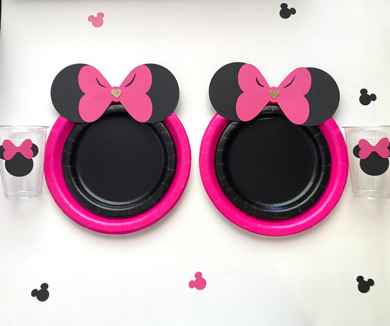 May include: Two black and pink paper plates with a pink bow and black ears on top. The plates are on a white surface with black and pink mouse ears scattered around.