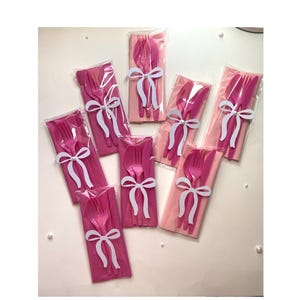 May include: Pink plastic cutlery sets, each including a fork, knife, and spoon, are packaged with a pink napkin and tied with a white bow. The sets are arranged on a white surface.