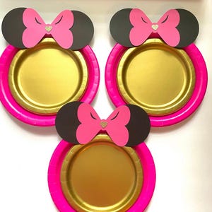 May include: Three gold paper plates with pink rims and black mouse ears with pink bows and gold glitter hearts.