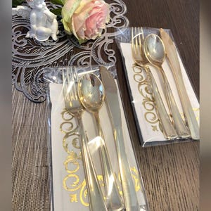 May include: Gold-colored disposable cutlery sets, including forks, spoons, and knives, packaged on decorative napkins. A white cherub statue and a pink rose are also visible.