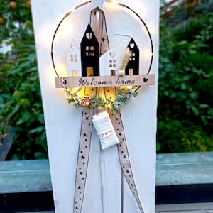 Può includere: Un cartello di benvenuto decorativo in legno con un design circolare. Il cartello presenta piccoli ritagli di case, una stella e uno striscione con le parole "Welcome Home". È adornato con luci fatate e nastri a forma di stella.