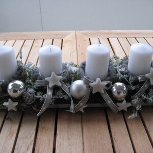 May include: A rectangular Advent candle holder with four white candles. The holder is decorated with gray ribbons, star-shaped ornaments, silver baubles, and greenery. The arrangement sits on a wooden table.