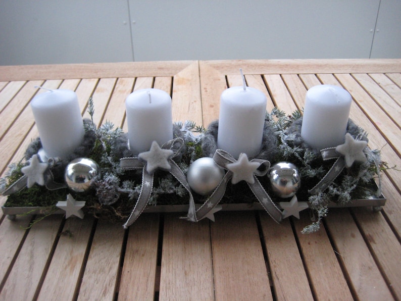 May include: A rectangular Advent candle holder with four white candles. The holder is decorated with gray ribbons, star-shaped ornaments, silver baubles, and greenery. The arrangement sits on a wooden table.