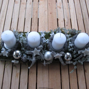 May include: A rectangular Advent candle holder with four white pillar candles. The candles are surrounded by gray felt, greenery, silver ornaments, and star-shaped decorations. Gray ribbons and a wooden surface complete the festive arrangement.