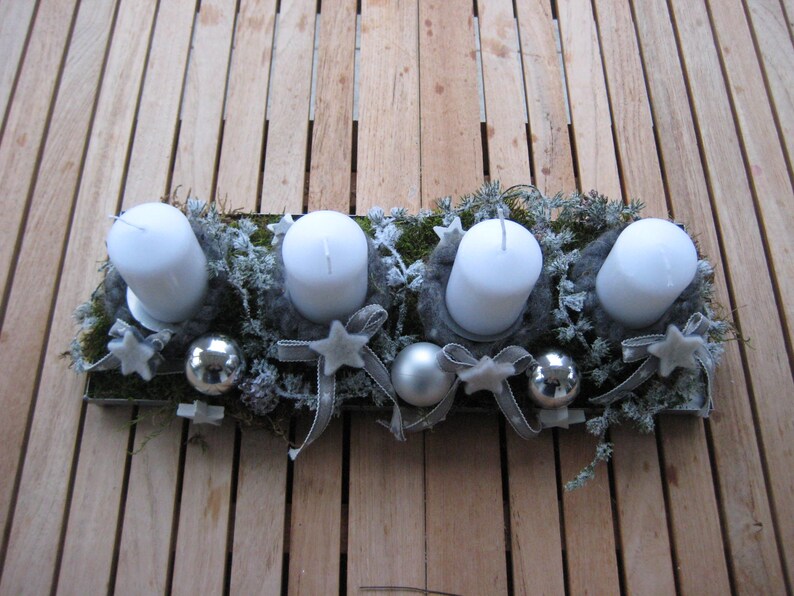 May include: A rectangular Advent candle holder with four white pillar candles. The candles are surrounded by gray felt, greenery, silver ornaments, and star-shaped decorations. Gray ribbons and a wooden surface complete the festive arrangement.