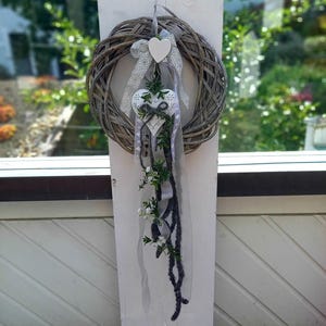 May include: A decorative wreath made of woven branches, adorned with white heart-shaped ornaments and ribbons. The wreath features a trailing arrangement of greenery and dark gray accents, creating a rustic and charming aesthetic. Perfect for home decor.