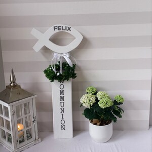 以下が含まれることがあります： 「FELIX」と書かれた白い木製の魚、白い木製のブロックに「COMMUNION」と書かれたもの、緑の葉のついた小さな白い花輪、緑の植物が入った白い鉢、そして中にろうそくが入った白いランタン。これらのアイテムは、2 つの引き出しのある白い棚の上に置かれています。