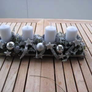 May include: A rectangular metal tray holds four white pillar candles, surrounded by greenery, silver ornaments, and star-shaped decorations. Gray ribbons and small stars add to the festive arrangement, perfect for holiday decor.