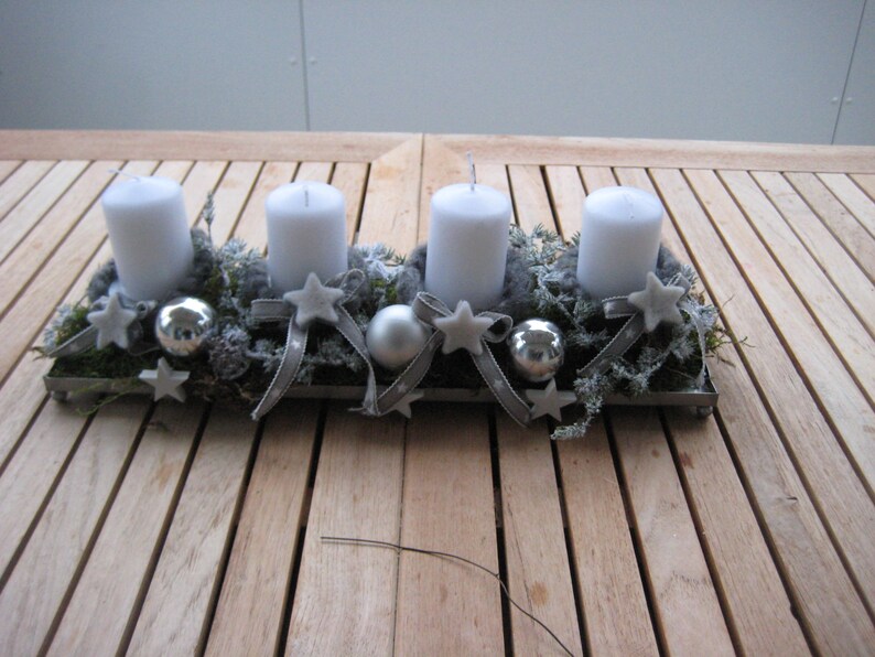 May include: A rectangular metal tray holds four white pillar candles, surrounded by greenery, silver ornaments, and star-shaped decorations. Gray ribbons and small stars add to the festive arrangement, perfect for holiday decor.