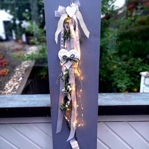 May include: A white, heart-shaped, decorative piece with a floral design hangs from a wooden post. The piece is adorned with white ribbon and a string of white fairy lights.
