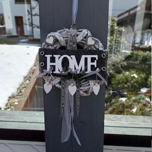 Può includere: Una ghirlanda circolare intrecciata fatta di rami grigio chiaro con una striscia di feltro grigio scuro al centro che riporta il testo bianco HOME. La ghirlanda è decorata con piccoli cuori in legno bianco, un nastro a quadri blu e bianco.