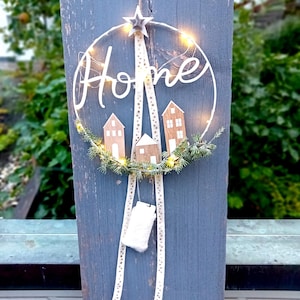 Może przedstawiać: Okrągły wieniec z napisem "Home" w kursywie, podświetlony małymi światełkami. Wieniec zawiera miniaturowe drewniane domki i jest ozdobiony zielenią i gwiazdą. Poniżej wisi wstążka z etykietą.