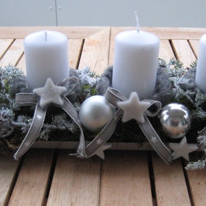 May include: A rectangular metal candle holder with four white pillar candles. The holder is decorated with silver ornaments, star-shaped accents, and gray ribbon. The arrangement sits on a wooden surface, creating a festive centerpiece.