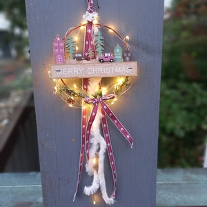 May include: A Christmas wreath with a wooden sign saying "Merry Christmas." It features miniature houses, trees, and cars, lit by warm white fairy lights. A red ribbon with white stars and feathers adds a decorative touch. The wreath is a festive decoration.