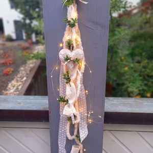 May include: A decorative hanging ornament featuring a cream-colored heart with embroidered details, adorned with faux greenery and fairy lights. The ornament is suspended by a ribbon and includes various textured ribbons and fabric elements.