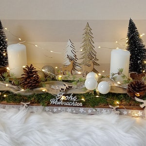 Könnte beinhalten: Ein festlicher Weihnachts-Tischschmuck mit vier weißen Kerzen, Miniatur-Weihnachtsbäumen, Tannenzapfen und dekorativen Holzelementen. Die Anordnung umfasst Lichterketten und den Text "Frohe Weihnachten".