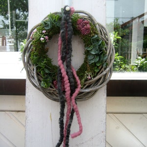 May include: A woven wreath with pink and black yarn hanging from it. The wreath is decorated with greenery and small pink flowers.