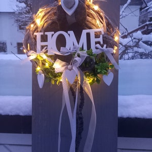 Puede incluir: Un letrero de madera blanco con la palabra "HOME" está unido a una corona de ramas marrones con luces blancas y cinta blanca. La corona está colgada en una superficie de madera gris.