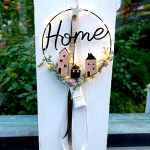 Puede incluir: Una corona circular con la palabra "Home" en escritura negra. La corona presenta casas de madera en miniatura, vegetación y pequeñas luces. Una estrella blanca y cintas cuelgan de la parte superior, creando un acento decorativo para el hogar.