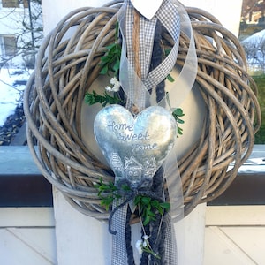 May include: A woven grapevine wreath with a heart-shaped metal ornament that reads "Home Sweet Home." The wreath is decorated with gray and white checkered ribbon, a white wooden heart, and greenery.