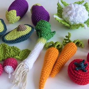 Pode incluir: Uma coleção de vegetais de crochê, incluindo um abacate verde, uma berinjela roxa, uma couve-flor branca, um alho-porro verde, duas cenouras laranja e três tomates vermelhos.
