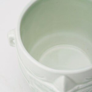 May include: A white ceramic planter with a textured, sculpted design. The planter has two handles on the sides and a wide, round opening.