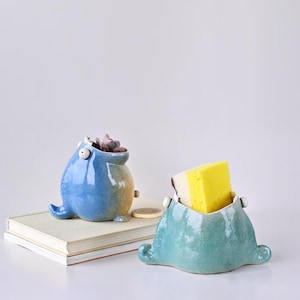 May include: Two whimsical ceramic containers. One is blue with a succulent inside, the other is teal and holds a yellow sponge. Both have a unique, cartoonish design with small details and a glossy finish. They sit on a stack of books.