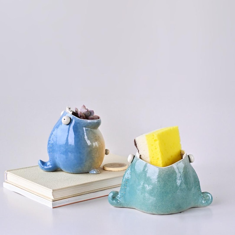 May include: Two whimsical ceramic containers. One is blue with a succulent inside, the other is teal and holds a yellow sponge. Both have a unique, cartoonish design with small details and a glossy finish. They sit on a stack of books.