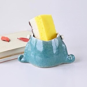 May include: A turquoise ceramic sponge holder with a yellow and white sponge inside. The holder has a unique shape with rounded edges and small decorative details. The background includes a book and some red leaves.