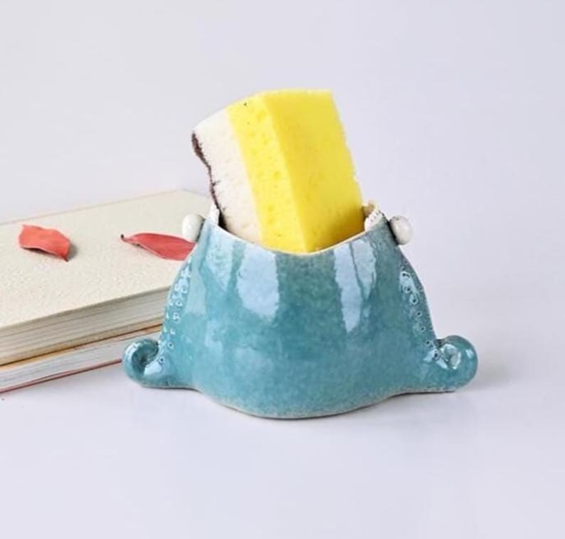 May include: A turquoise ceramic sponge holder with a yellow and white sponge inside. The holder has a unique shape with rounded edges and small decorative details. The background includes a book and some red leaves.